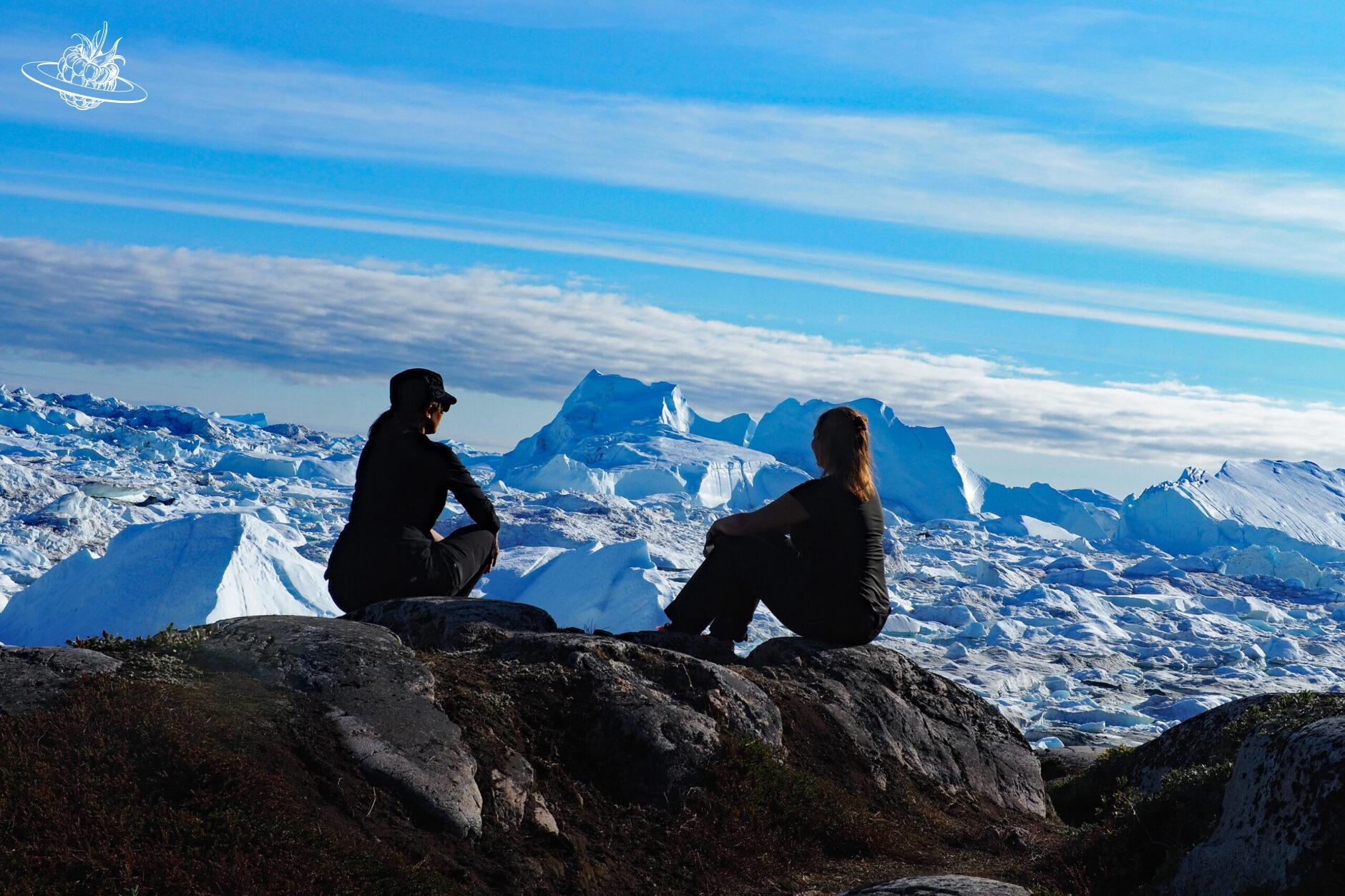 zwei Frauen sitzend auf Felsen vor einer riesigen Eisberglandschaft