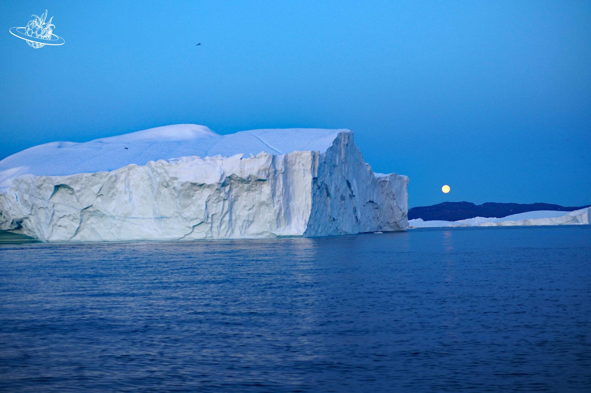 grosser Eisberg und im Hintergrund der Vollmond, welcher über das Meer scheint