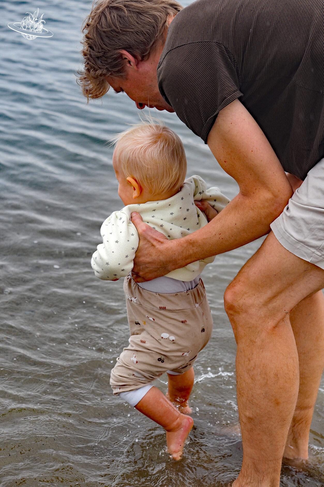 Mann trägt Kind so, dass dessen Füsse das Wasser berühren