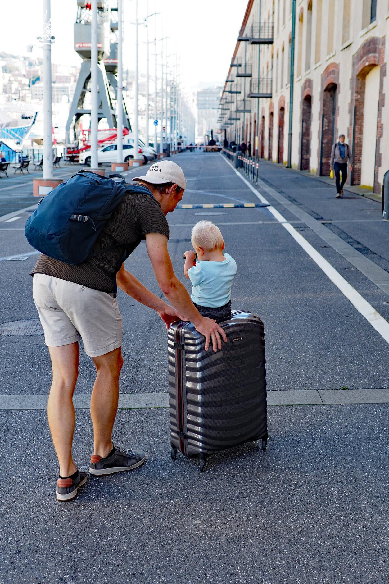 Mann schiebt Koffer, auf welchem ein Kind sitzt